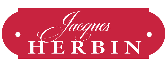 A red, oval-shaped label with two small holes on each side. The label displays the words Jacques HERBIN in elegant white script and capital letters, reflecting the brand’s heritage in kalligrafie and exquisite schreibtinte.